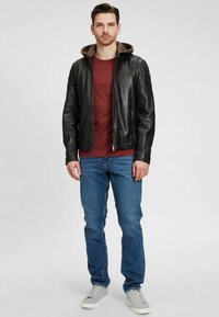 Chaqueta de cuero negra con capucha marrón, cierre de cremallera y detalles acanalados, combinada con una camisa burdeos y jeans azules, complementada con zapatillas grises.