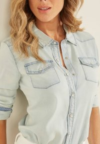 Cămașă din denim albastru deschis cu mâneci lungi suflecate. Are două buzunare pe piept și nasturi prindere din metal argintiu pe toată lungimea închiderii.