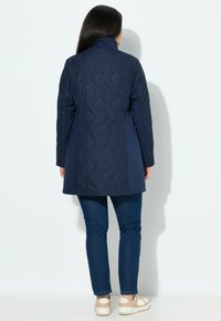 Kvinna med långt mörkt hår iklädd en marinblå vadderad jacka, blå jeans och beigevita sneakers som står mot en enkel vit bakgrund.