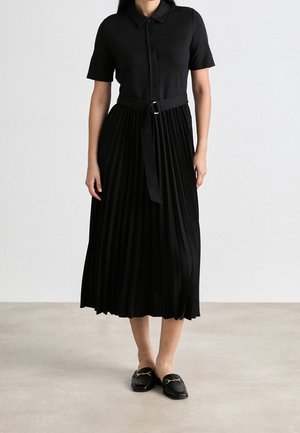 Kobieta ubrana w czarną sukienkę z krótkim rękawem, kołnierzykiem, plisowaną spódnicą, w talii przewiązaną paskiem oraz czarne wsuwane mokasyny z ozdobą w postaci złotego łańcuszka.