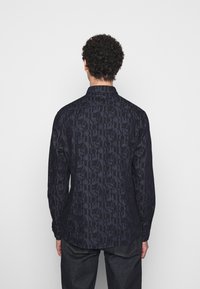Camicia a maniche lunghe in tessuto di un blu navy scuro, caratterizzata da un motivo testurizzato di grandi lettere ripetute. Colletto classico e orlo dritto.