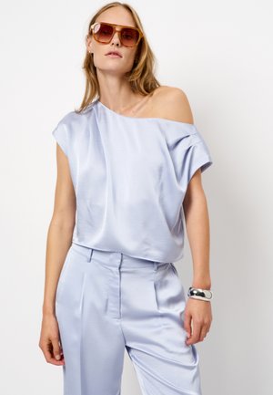 Lichtblauw satijnen top, off-shoulder ontwerp, brede mouwen, gecombineerd met bijpassende satijnen broek, vergezeld van een glanzende zilveren armband en oversized zonnebril.