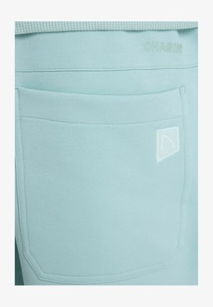 Hellblauer Stoff in Nahaufnahme, der eine große aufgenähte Tasche mit einem kleinen dreieckigen Logo und dem geprägten Schriftzug "CHASIN" nahe dem Bund zeigt.