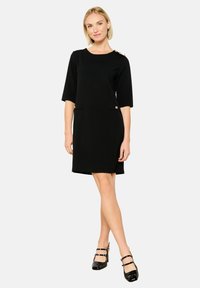 Vestido negro de manga corta con bolsillos laterales, cuello redondo y acentos de botones dorados en el hombro, hecho de una tela suave.