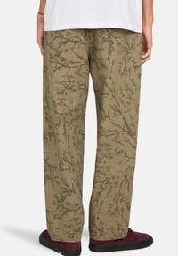 Pantalon kaki avec un motif ramifié en brun foncé, en coton, avec une coupe ample et des poches arrière. Agrémenté de baskets noires.