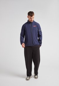 Chaqueta ligera de la marina con cuello alto, cremallera frontal y puños elásticos. Cuenta con un bolsillo con cremallera en la manga y un acento de logo rojo.
