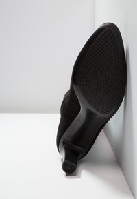 Chaussure à talon haut en daim noir avec une semelle en caoutchouc texturée, un bout pointu et un talon lisse. Présente un design minimaliste sur un fond clair.