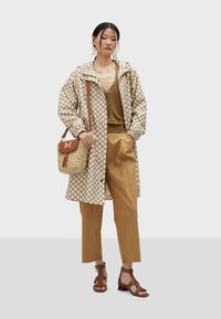 Lekki, beżowy płaszcz przeciwdeszczowy w białe kropki, w połączeniu z brązową bluzką i beżowymi spodniami. Dodatki to pleciona torba i brązowe sandały.
