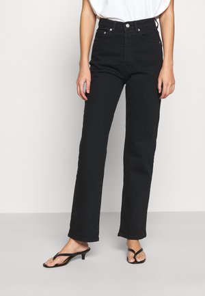 Osoba ubrana w czarne, proste jeansy z wysokim stanem, czarne sandały na obcasie z paskiem między palcami oraz biały top, stojąca na jednolitym tle.