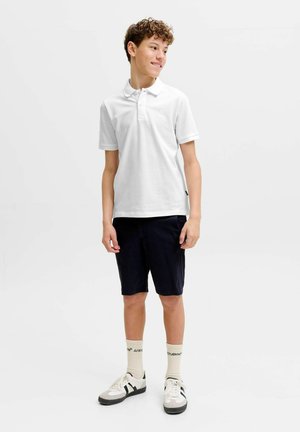 Tenåringsgutt med krøllete hår som har på seg hvit poloskjorte, svarte shorts, hvite sokker og hvite joggesko, står og ser til siden.