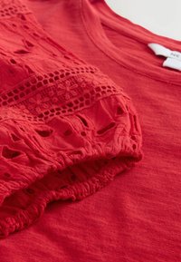 Chemise rouge à manches courtes avec détails en dentelle et découpes sur la manche bouffante, vue en gros plan avec poignet élastique froncé.