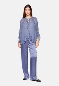 Flowy lavender blouse with ruffled edges and a tie neckline, paired with shiny, wide-leg lavender trousers and black flat shoes.