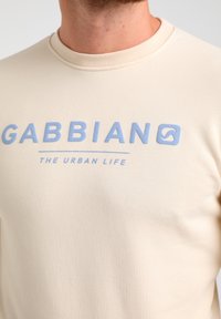 Sudadera de un beige claro con cuello redondo, que presenta texto bordado en azul "GABBIAN" y "LA VIDA URBANA". Textura suave, diseño minimalista.