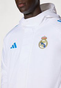 Hvid atletisk jakke med hætte, udstyret med blå Adidas striber og et Real Madrid-logo på brystet. Glat, letvægtsmateriale.