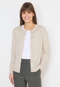 Femme souriante portant un cardigan beige boutonné sur une chemise blanche et un pantalon gris à cordon, debout devant un fond blanc.