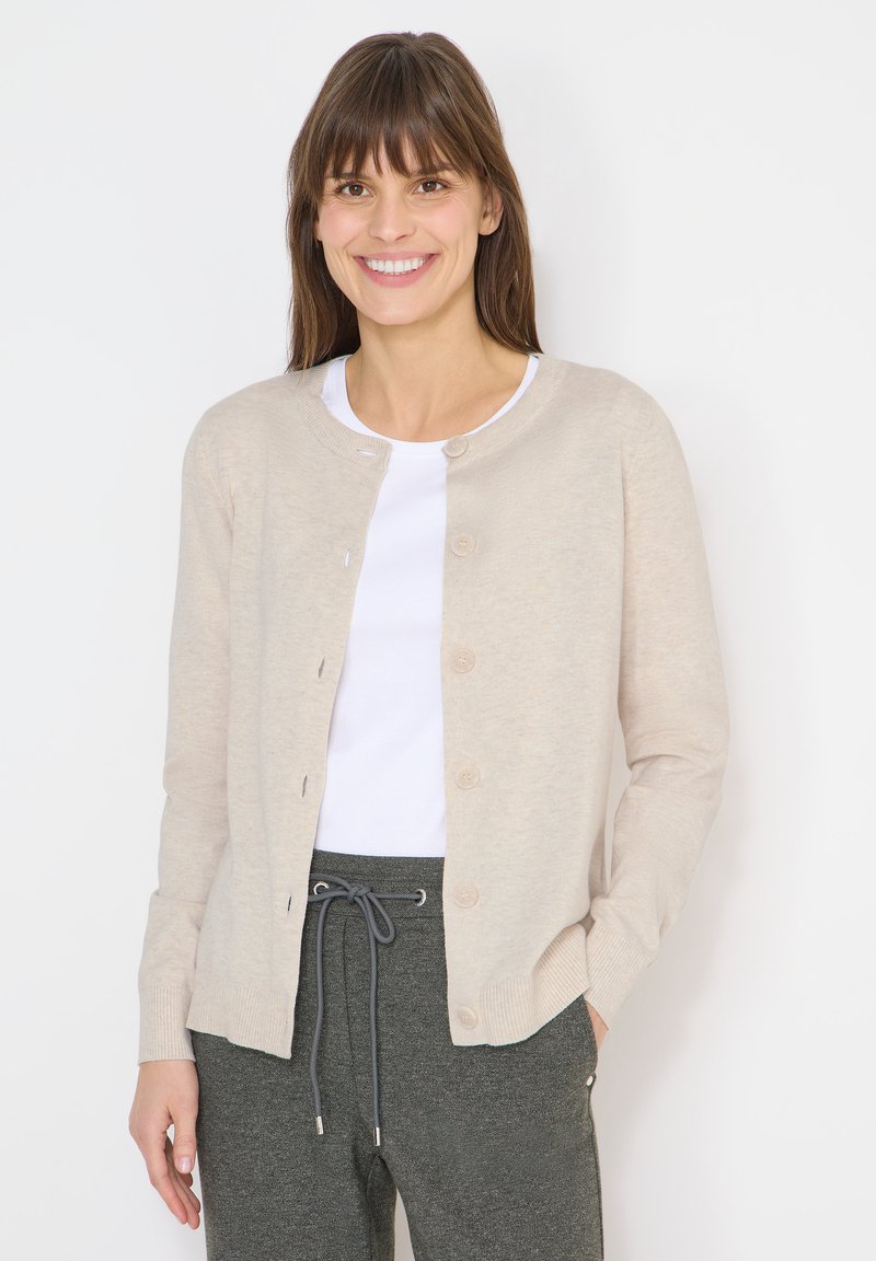 Femme souriante portant un cardigan beige boutonné sur une chemise blanche et un pantalon gris à cordon, debout devant un fond blanc.