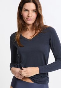 Mujer con el cabello largo y castaño, vistiendo una camiseta de manga larga azul marino con escote en V, ajustando el dobladillo, sobre un fondo blanco.