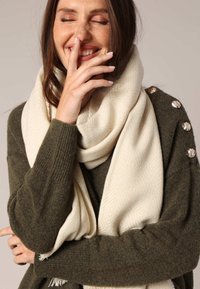 Écharpe crème avec texture en chevrons drapée autour d'une personne portant un pull vert foncé avec des boutons métalliques décoratifs à l'épaule.