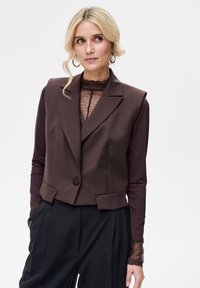 Brązowa, bezrękawnikowa, dopasowana kamizelka z pojedynczym guzikiem, nałożona na ciemnobrązową, długorękawową koronkową bluzkę, zestawiona z czarnymi spodniami.