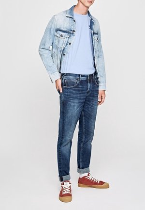 Homme portant une veste en jean bleu clair, un t-shirt bleu clair, un jean bleu foncé retroussé, des chaussettes blanches et des baskets rouges, debout avec une main dans la poche.