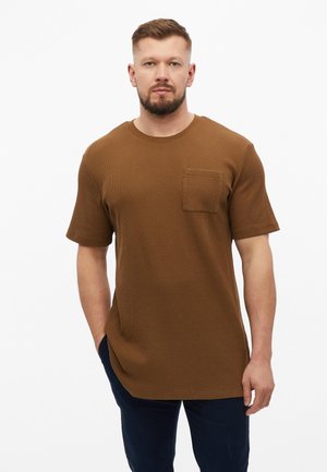 Mann trägt ein braunes kurzärmeliges, strukturiertes Hemd mit Brusttasche und dunkle Hose, steht vor einem schlichten weißen Hintergrund.
