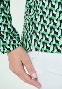 Camicia a motivi geometrici verdi con accenti neri e bianchi, caratterizzata da polsini arricciati e spacchi laterali, abbinata a pantaloni bianchi con dettagli di borchie.