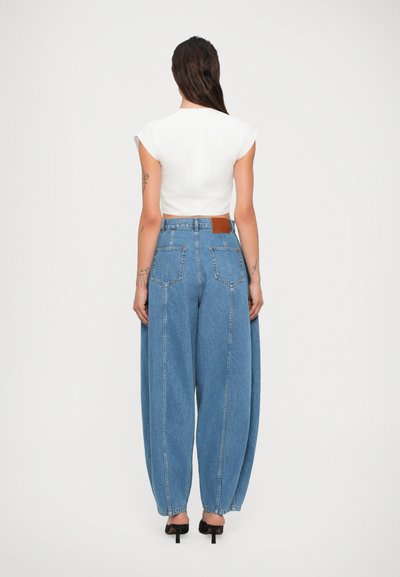 Femme se tenant de dos, portant un crop top blanc, un jean large taille haute bleu, des chaussures à talons noires et montrant des tatouages sur les bras.