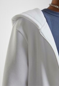Felpa bianca con zip, caratterizzata da una texture morbida, cappuccio a doppio strato e dettagli in metallo sulla zip, indossata su una maglietta blu a coste.