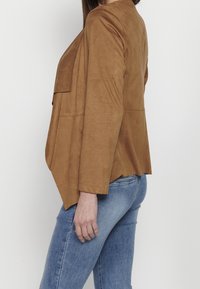 Femme en vue latérale portant une veste en daim marron et un jean en denim bleu sur un fond blanc uni.