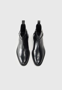 Botas de tobillo de cuero negro con acabado liso, punta afilada, cremallera lateral y tira de agarre. Diseño elegante con acentos mínimos.