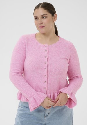 Woman wearing a light pink buttoned cardigan with ruffled cuffs, paired with light blue jeans, standing against a plain white background.