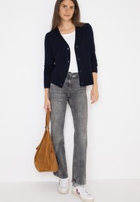 Marinblå stickad cardigan med långa ärmar och knappar, vit t-shirt, grå utsvängda jeans och brun mockaväska; modellen har på sig vita sneakers.