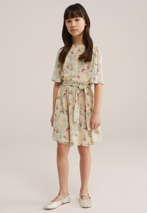 Noor tüdruk, kellel on pikad pruunid juuksed ja kes kannab kreemjat lillemustrilist põlvikku pikkusega kleiti vöökohal paelaga ning valgeid sädelevaid Mary Jane stiilis kingi, seismas.