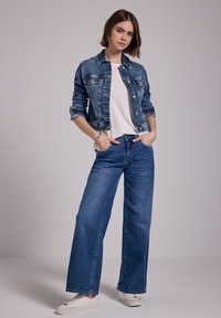 Giacca di jeans con chiusura a bottoni, di colore azzurro chiaro, e due tasche sul petto, abbinata a jeans a gamba larga blu e sneakers bianche.