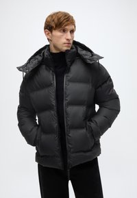 Μαύρο μπουφάν τύπου puffer με κουκούλα, με σχεδιασμό με ραφές, μπροστινό φερμουάρ και δύο πλαϊνές τσέπες. Λείο, μονωτικό υλικό.
