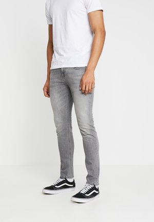 Homme portant un jean slim gris clair, des baskets noires et blanches, et un t-shirt blanc uni à manches courtes, debout sur un fond blanc.