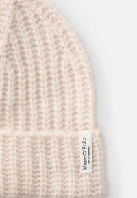 Bonnet beige en tricot fabriqué en matériau doux, avec un revers plié et une petite étiquette blanche portant le texte "Marc O'Polo" sur le côté.