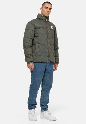 Grønn isolert pufferjakke med høy krage, glidelås, sydd detaljer og logo på brystet; kombinert med blå denim jeans og hvite joggesko.