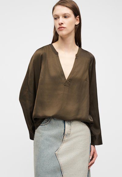 Γυναίκα που φοράει μια άνετη καφέ μπλούζα με V λαιμόκοψη και μακριά μανίκια, μαζί με μια ψηλόμεση δίχρωμη τζιν φούστα, μπροστά σε απλό φόντο.