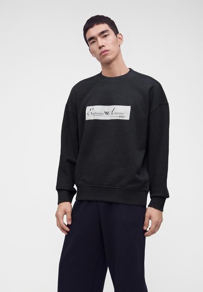 Млад мъж, облечен в черен суичър с логото на "Emporio Armani" на гърдите и широки тъмни панталони, стои на светъл едноцветен фон.
