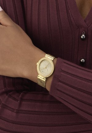 Montre en or avec un cadran rond orné de cristaux et un bracelet à motifs, portée au poignet. Le pull a une texture en côtes de couleur bourgogne.