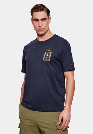 Uomo che indossa una T-shirt blu navy con stemma ricamato e pantaloni cargo verde oliva, in piedi contro uno sfondo chiaro e uniforme.