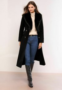 Abrigo negro de doble botonadura con cuello de piel sintética, combinado con un suéter de punto trenzado color crema y jeans azules, complementado con botas negras hasta la rodilla.