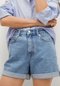 Shorts en denim bleu avec revers roulés, dotés d'un design à cinq poches, d'une braguette zippée et d'une texture délavée claire. Associés à une chemise à rayures bleues.