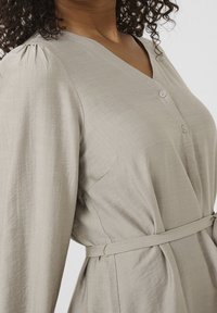 Femme portant une robe beige clair boutonnée avec des manches longues bouffantes et une fine ceinture nouée à la taille.