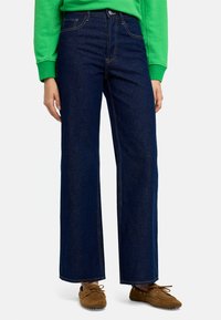 Niebieskie szerokie dżinsy z denimu z widocznym przeszyciem i zapięciem na guzik, połączone z brązowymi zamszowymi butami. Brak wzorów lub akcentów.