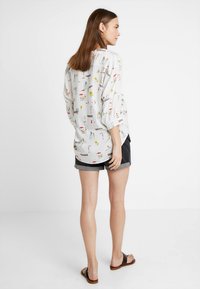 Blusa de cor clara, com padrões e gráficos divertidos de tema de verão, com um corte solto, mangas longas e detalhes de botões, combinada com calções cinzentos.