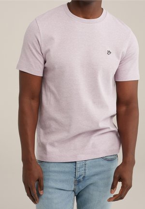 Homme portant un t-shirt à manches courtes violet clair avec un petit emblème de main sur la poitrine et un jean bleu clair, debout devant un fond uni.