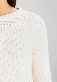 Nahaufnahme einer Person in einem weißen Strickpullover mit diagonalen Zopfmuster an der Schulter und dem Ärmel vor einem schlichten Hintergrund.
