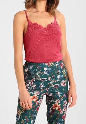 Woman wearing a sleeveless pink lace-trimmed top and high-waisted green floral pants, standing against a plain background.